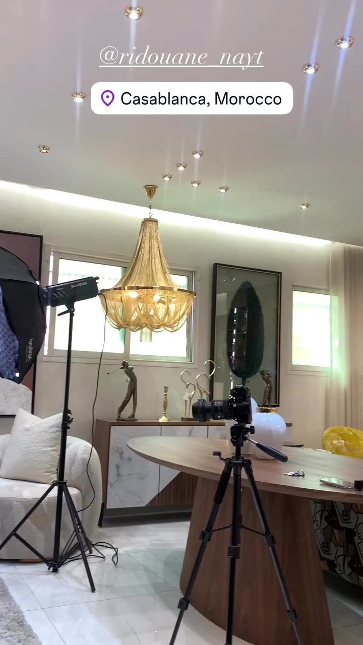 Salon meublé en cours de tournage : trépied caméra au premier plan et projecteur sur pied allumé à gauche.